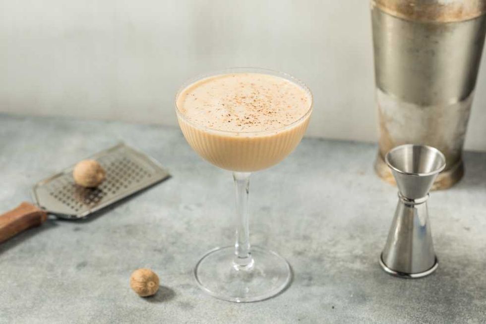 Creamy cocktail in a glass, with nutmeg, grater, and shaker on a gray countertop.