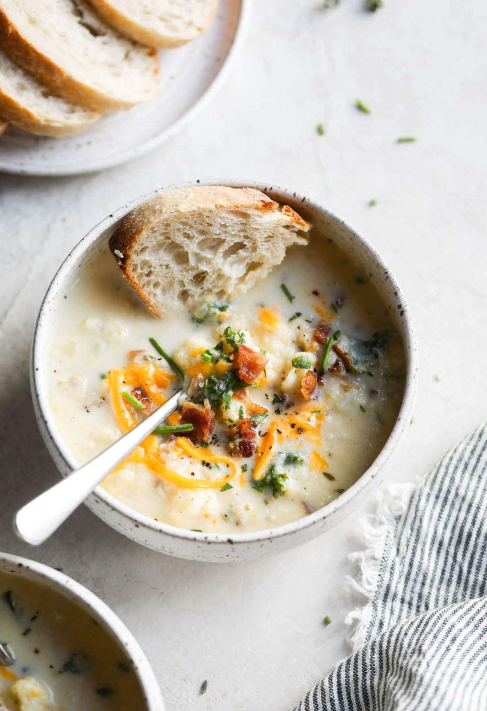 Creamy soup with bacon, cheese, herbs, and bread in a bowl on a white surface.