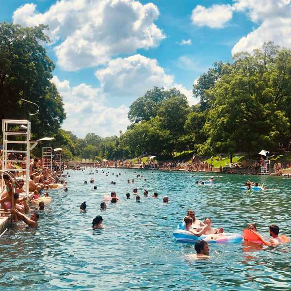 Crowded outdoor pool with swimmers, trees, and a bright blue sky with clouds.