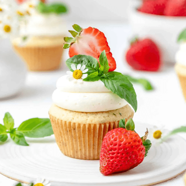 Cupcake with cream frosting, strawberry slice, mint, and small flower garnish on a white plate.