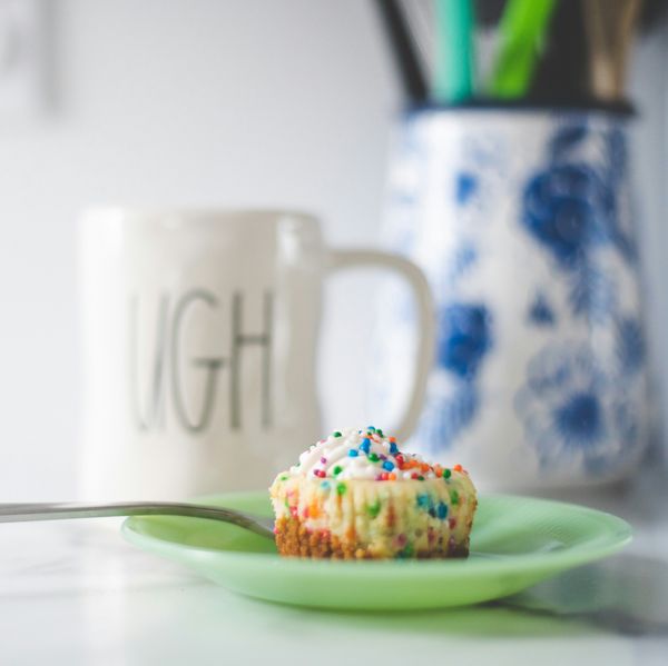 Cupcake with sprinkles on a green plate, spoon beside it, mug and vase in the background.