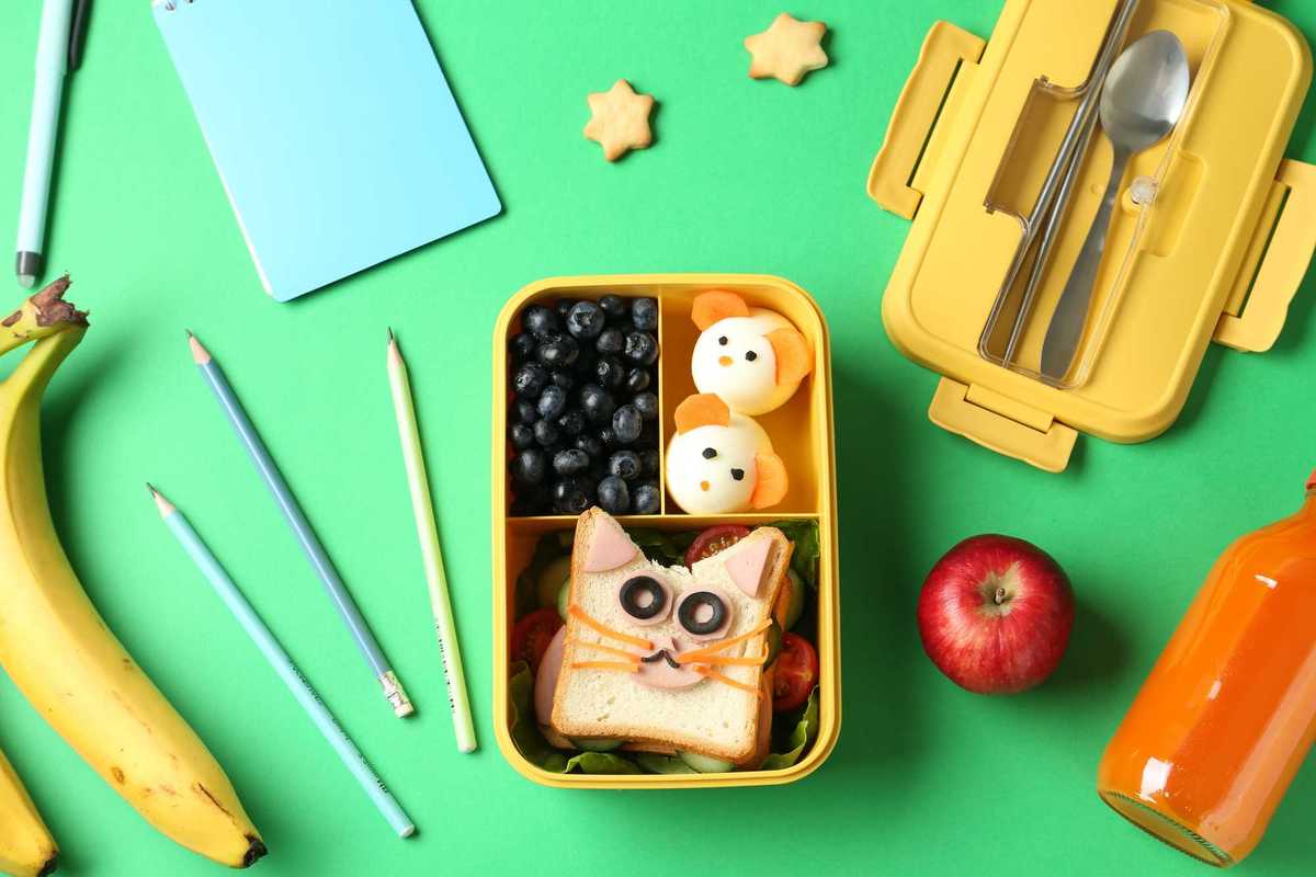 Cute animal-themed lunchbox with fruits, pencils, and juice on a green background.