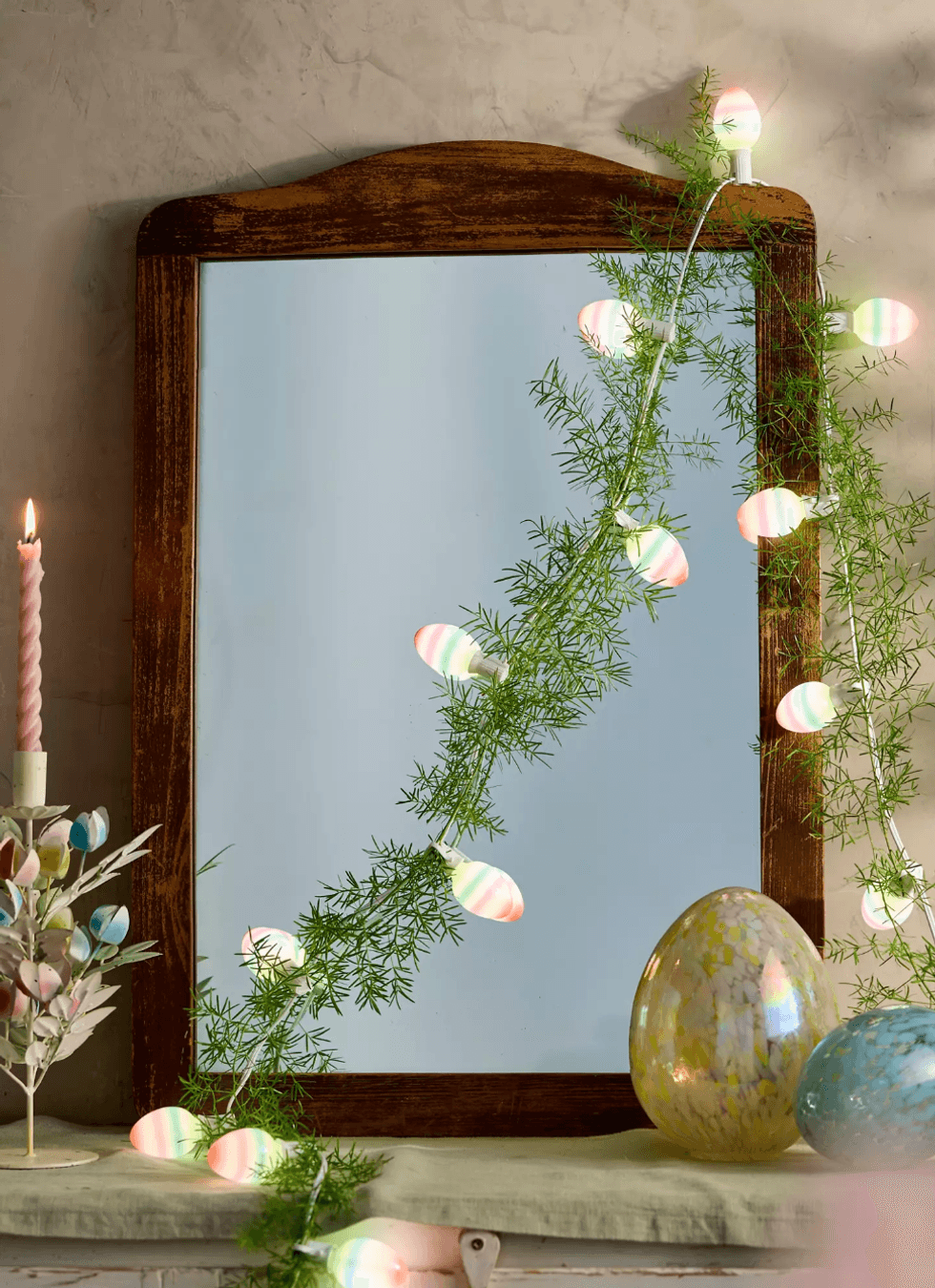 Decorated mirror with egg-shaped lights, leafy garland, candle, and colorful eggs.