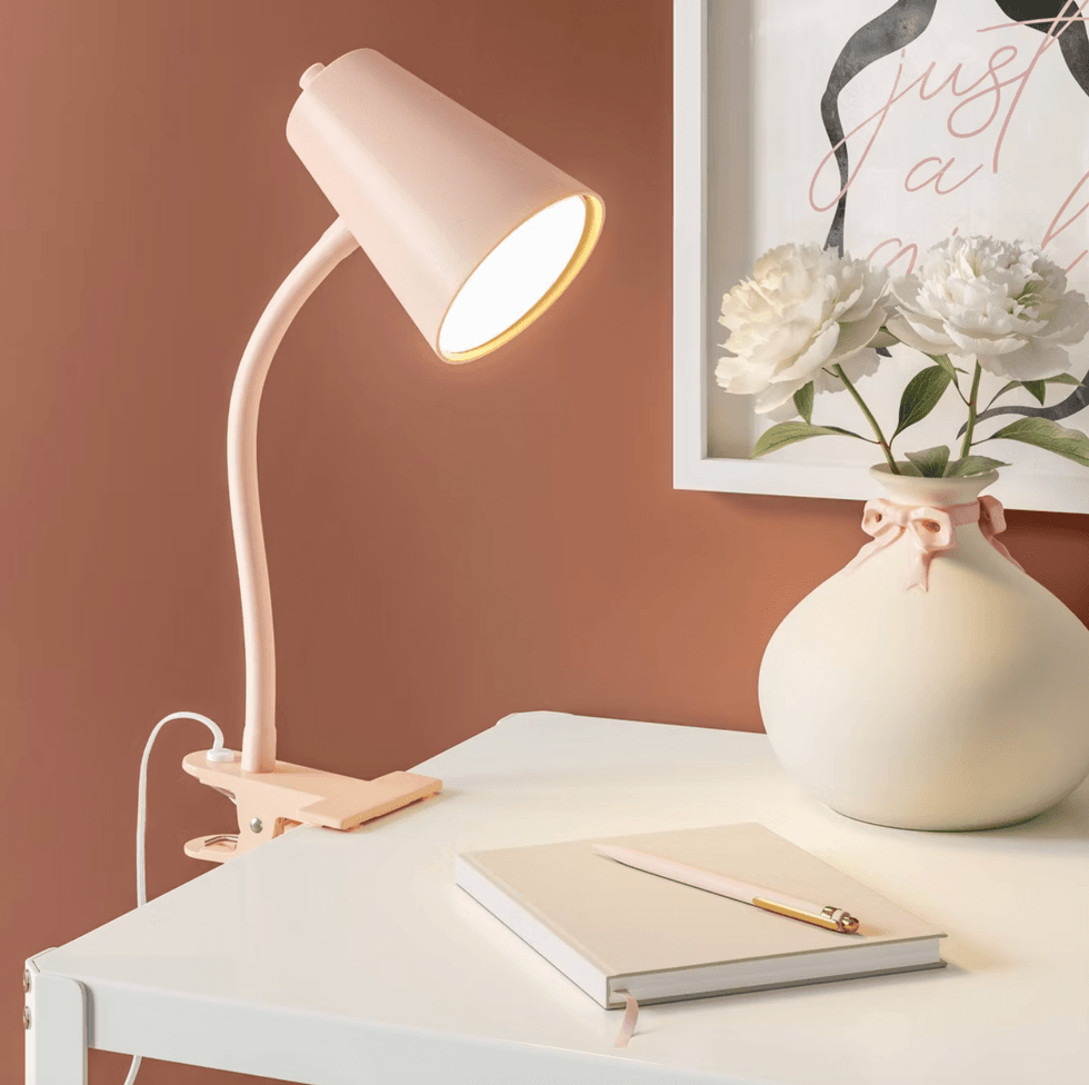 Desk with a pink lamp, notebook, pencil, and vase of flowers on a white table.