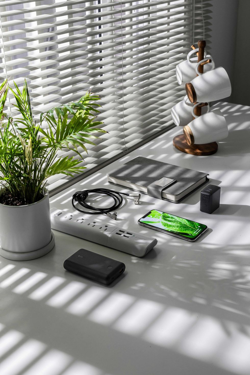 Desk with plant, smartphone, notebooks, power strip, and mug stand under window blinds.