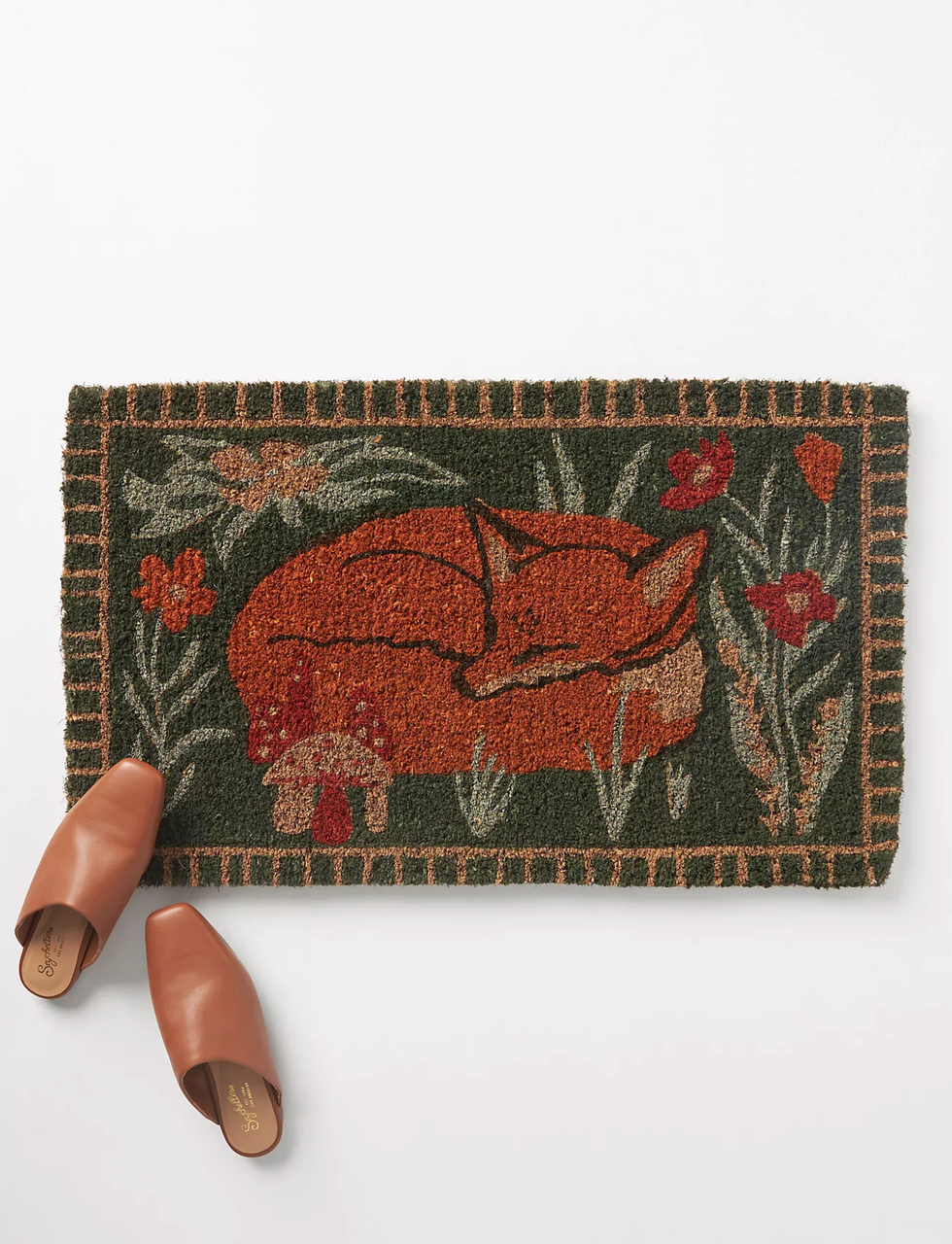 Doormat with a sleeping fox and flowers, next to brown slip-on shoes.