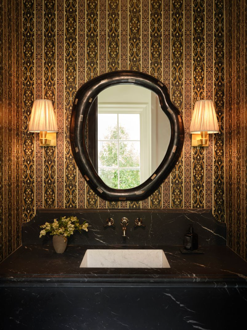 Elegant bathroom with ornate wallpaper, unique mirror, and wall sconces above a black sink.