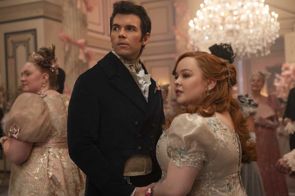 Elegant couple in period costumes at a lavish ballroom event, surrounded by other guests.