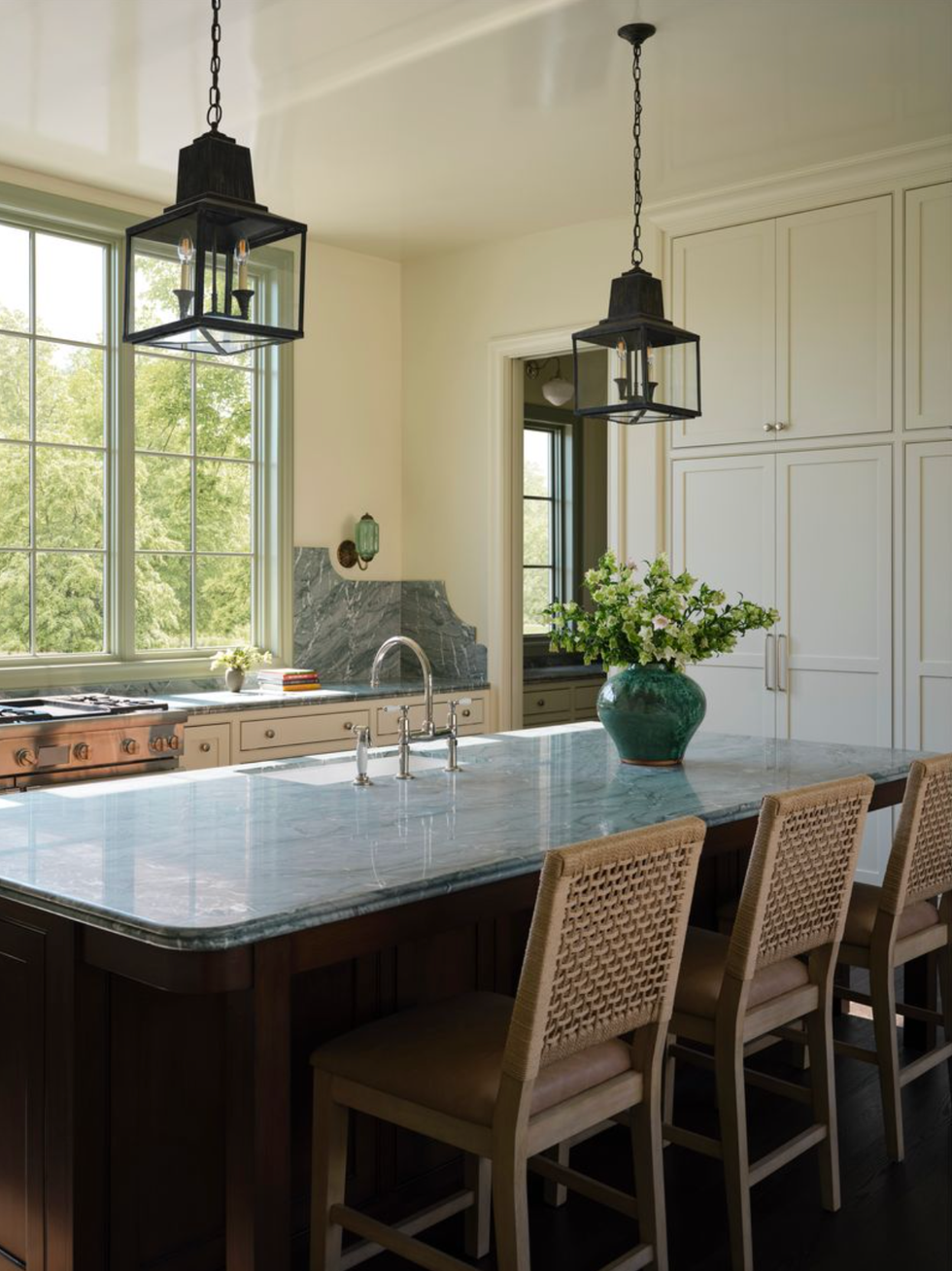 Elegant kitchen with marble island, woven chairs, and pendant lights over a scenic window view.