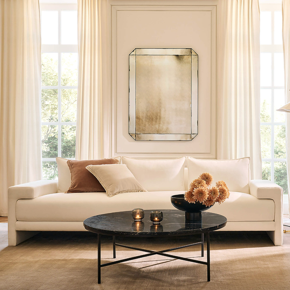 Elegant living room with a white sofa, round table, and large mirror, bathed in natural light.