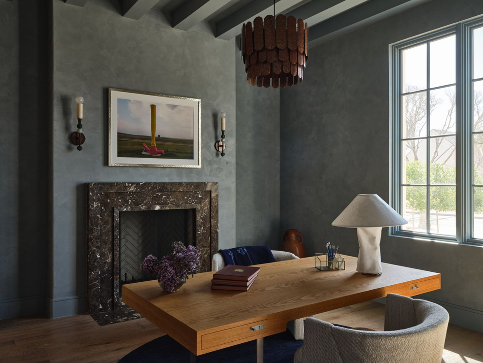 Elegant study with wooden desk, modern lamp, and a marble fireplace under natural light.