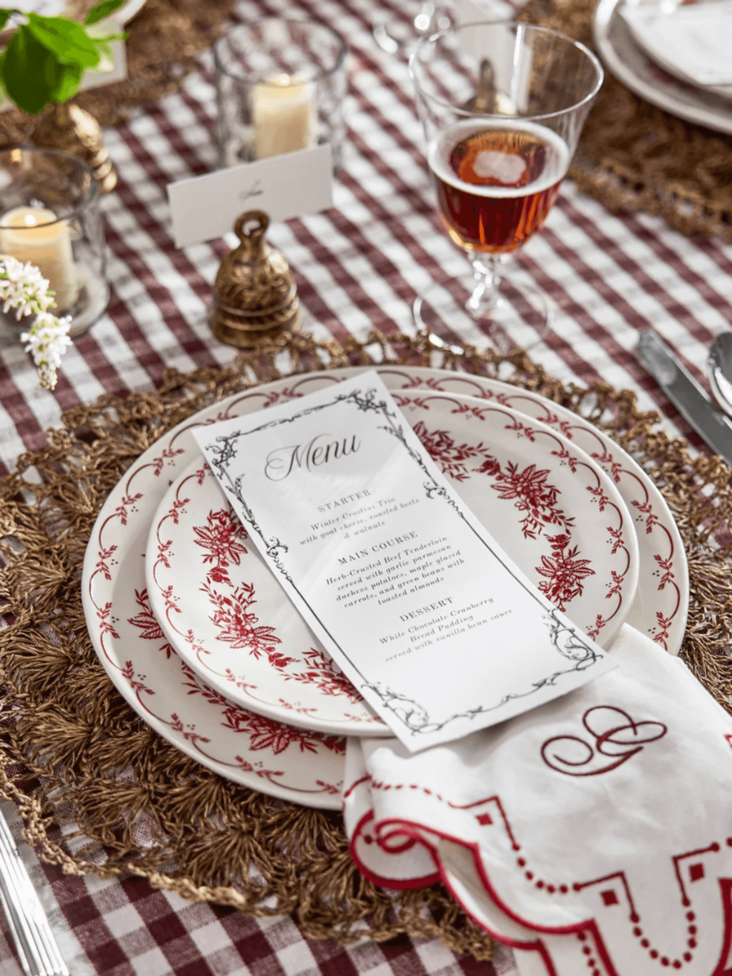 Elegant table setting with red patterned plates, menu, wine glass, and candle on checkered cloth.