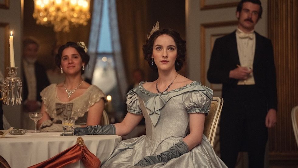 Elegant women in period dresses sit at a candlelit table in a grand room.