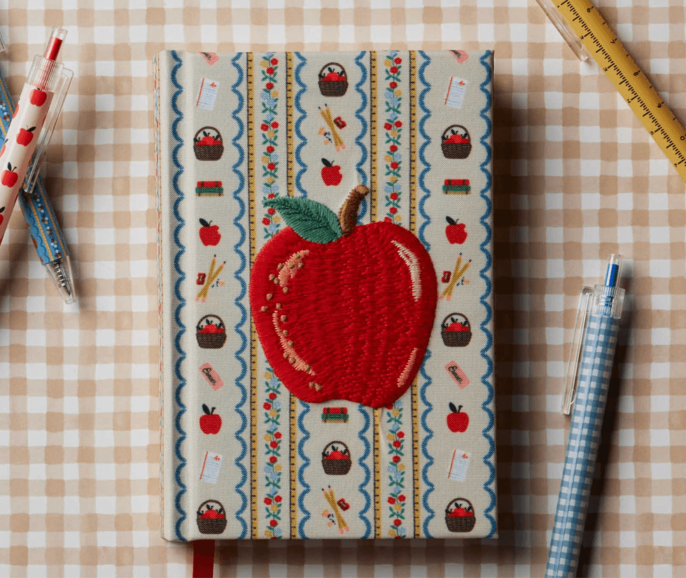 Embroidered apple notebook on checkered table, surrounded by pens and a ruler.