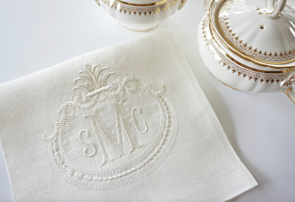 Embroidered white napkin with monogram beside ornate teapot and sugar bowl.