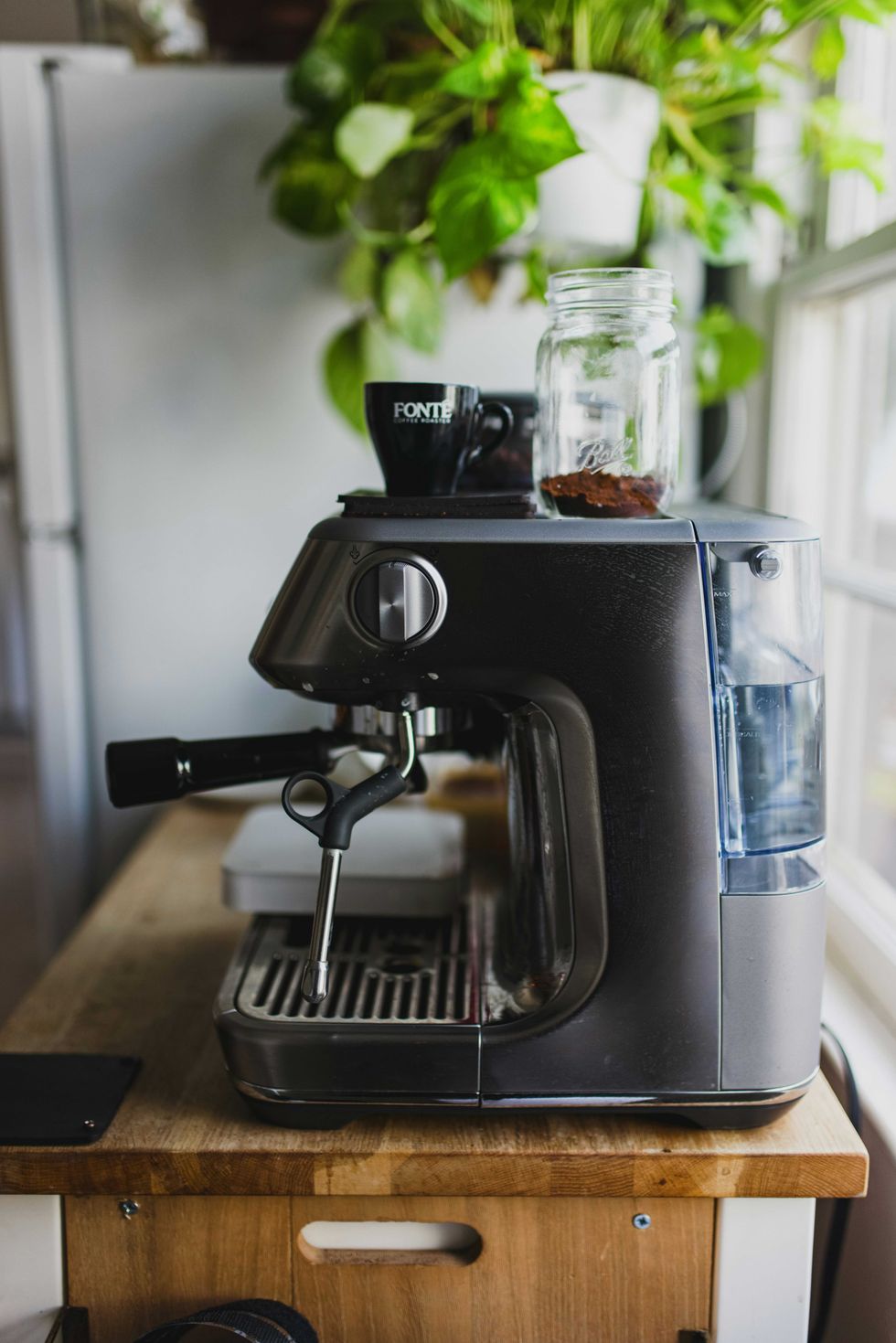 Espresso machine with a cup and jar atop, set on a wooden countertop near plants.