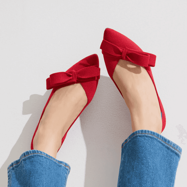 Feet in blue jeans wearing bright red ballet flats with bows against a white wall.