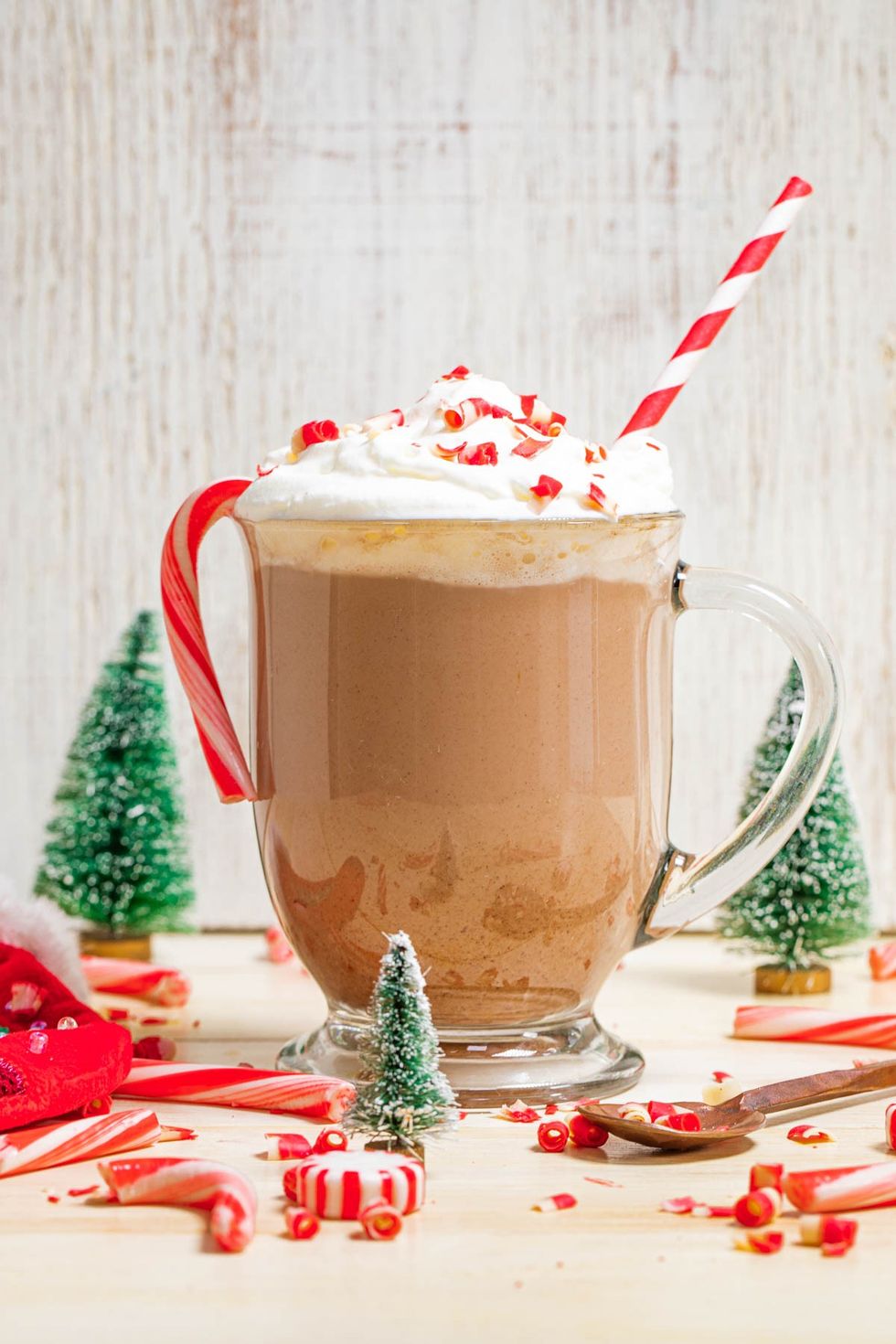 Festive hot chocolate with whipped cream, candy cane, and straw; tiny Christmas trees nearby.