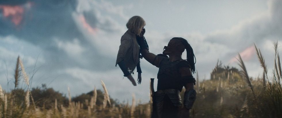 Figure holding a person in a grassy field under a cloudy sky.