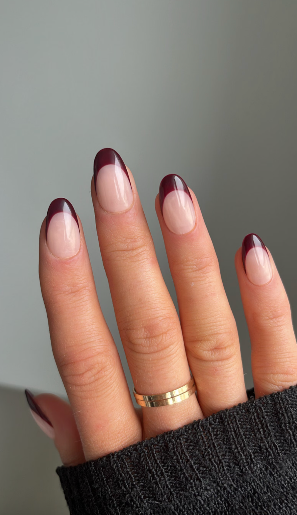 Fingers with dark burgundy French tip nails and a gold ring, against a gray background.