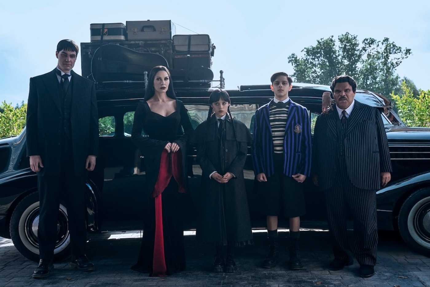 Five people in dark, formal clothing standing in front of a classic car with luggage on top Wednesday episodes