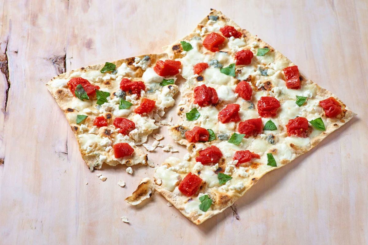 Flatbread pizza with tomatoes, cheese, and basil on a light wooden surface.