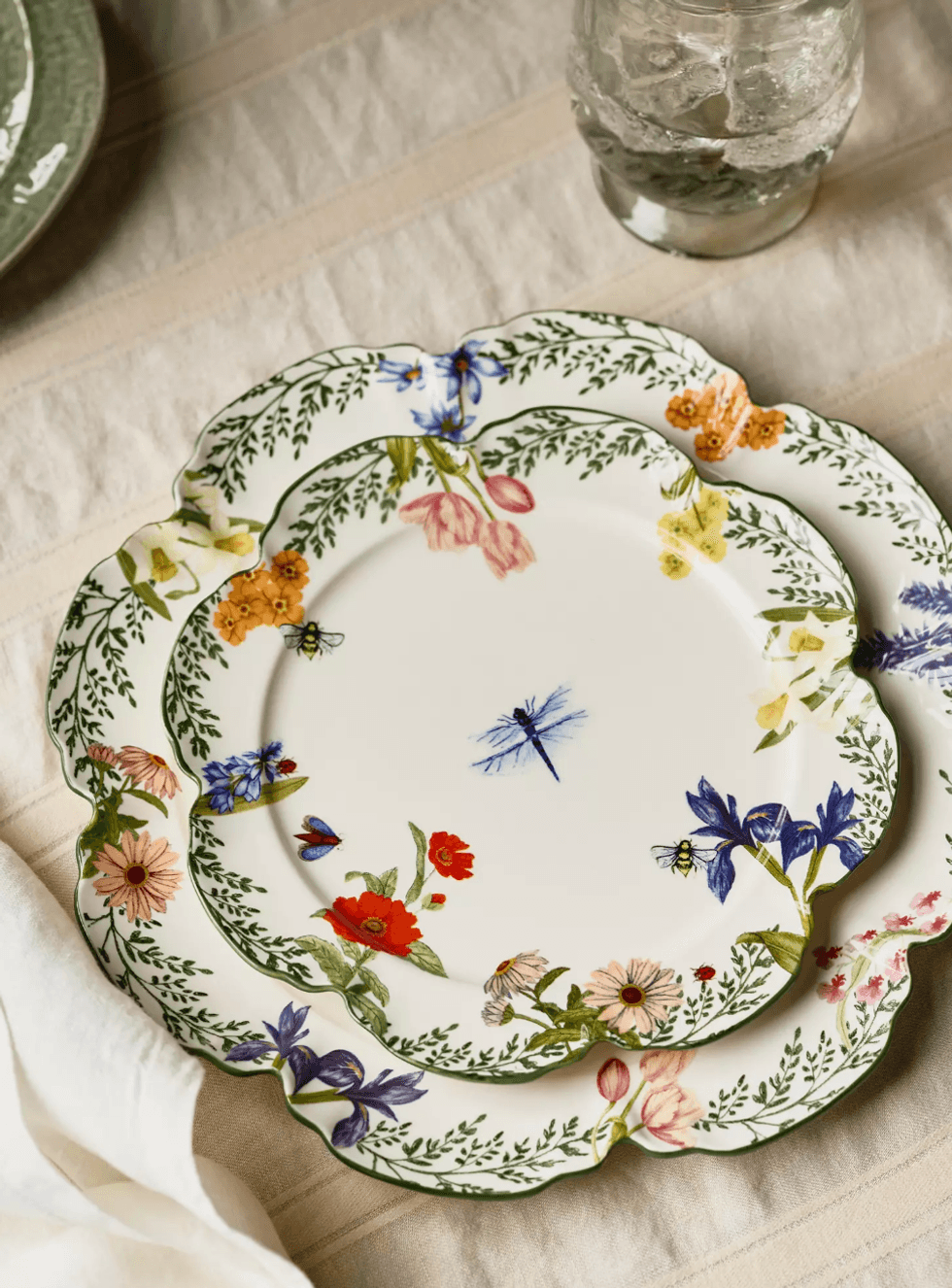 Floral and dragonfly patterned plates on a beige tablecloth with a glass nearby.