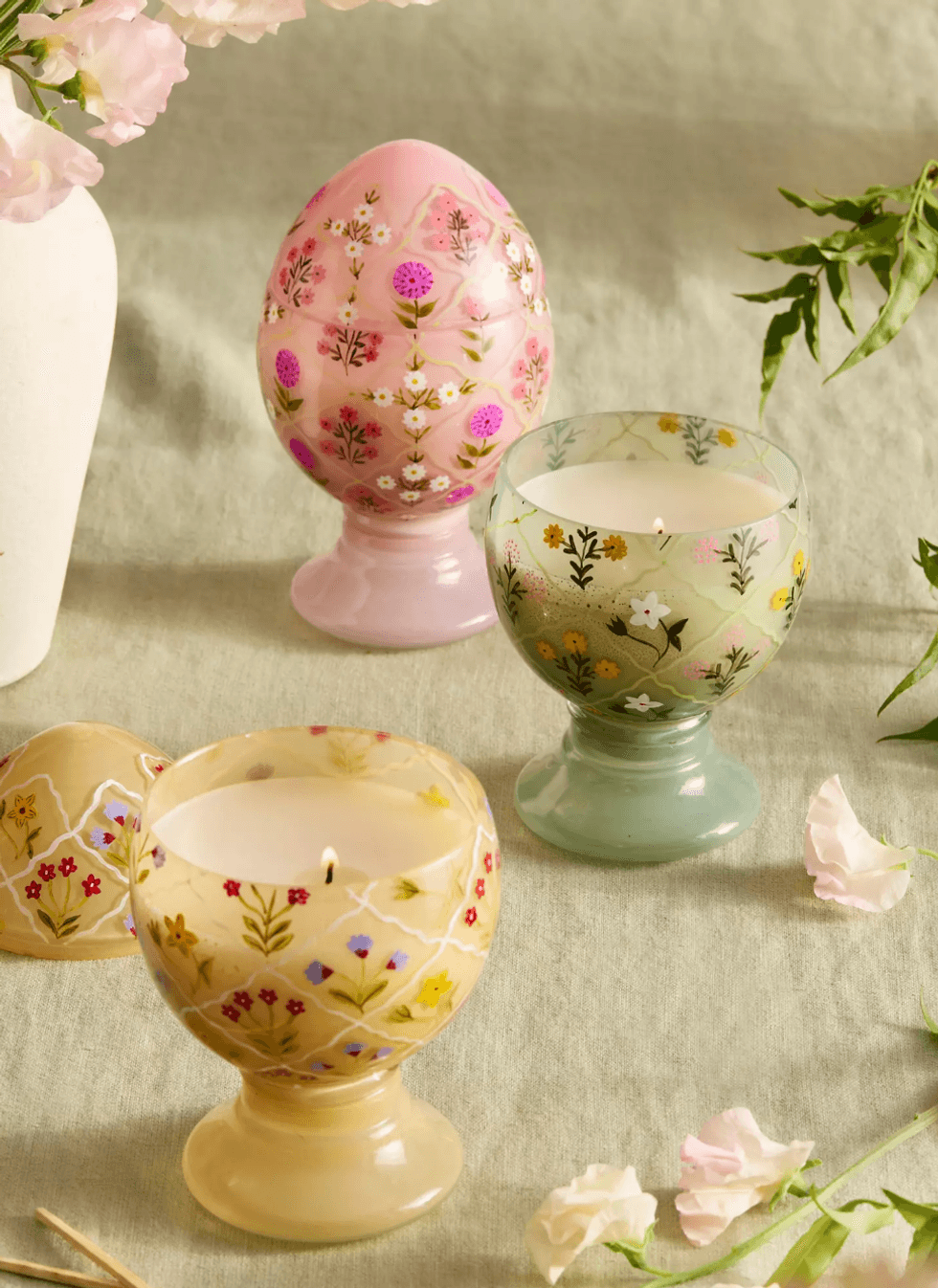 Floral egg and cup-shaped candles on a linen table with scattered petals.