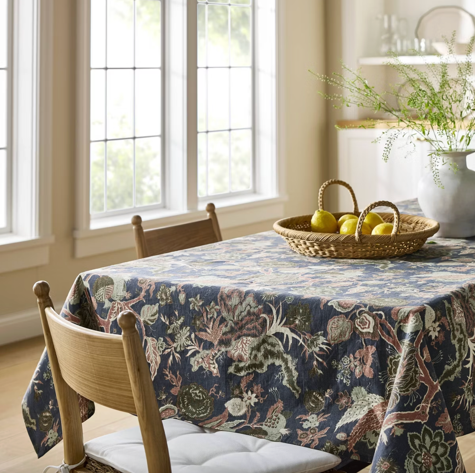 Floral tablecloth, basket of lemons, and vase on a dining table by bright windows.