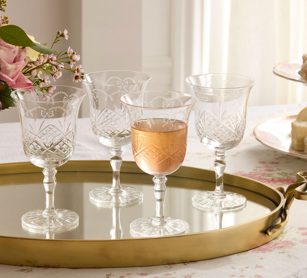 Four crystal glasses, one filled with ros\u00e9, on a mirrored tray with floral decor.