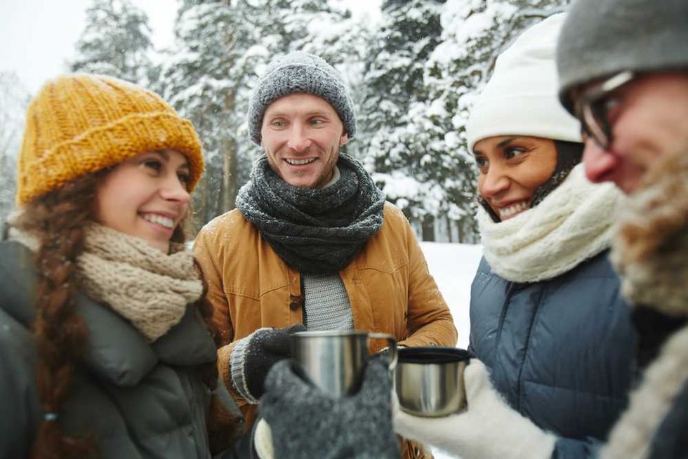 Four friends in winter attire enjoy hot drinks in a snowy forest setting.
