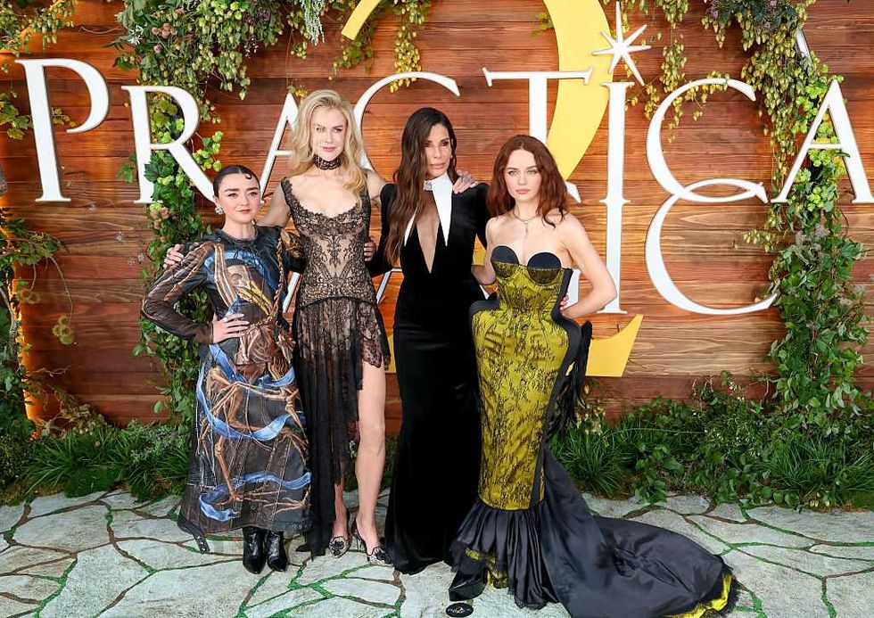 Four people in elegant dresses pose at an outdoor event, with greenery and a wooden backdrop.