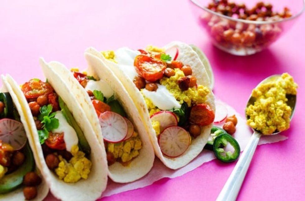 Four vegan breakfast tacos sit on a napkin on a pink surface.