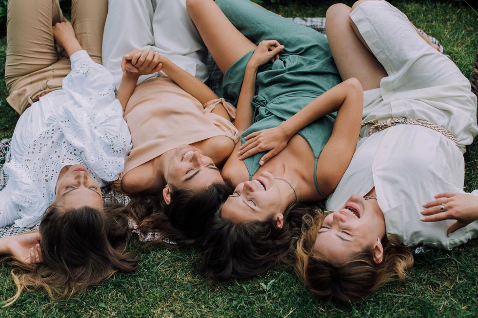 Four women lying on grass, laughing and wearing summer dresses.