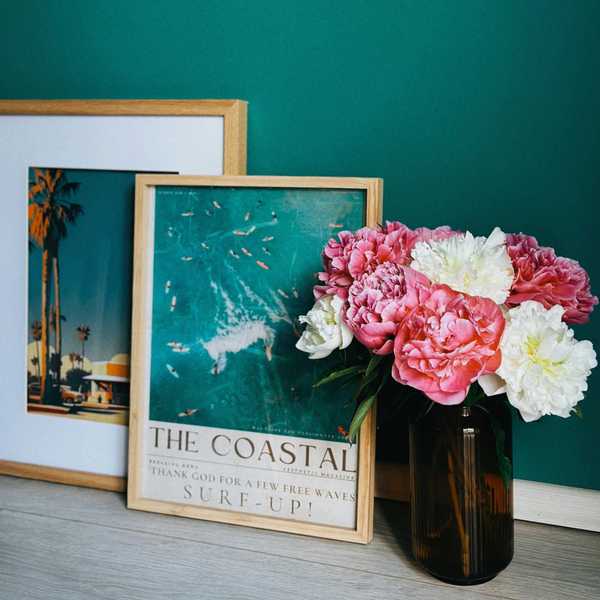 Framed beach posters and a vase with pink and white flowers against a teal wall.