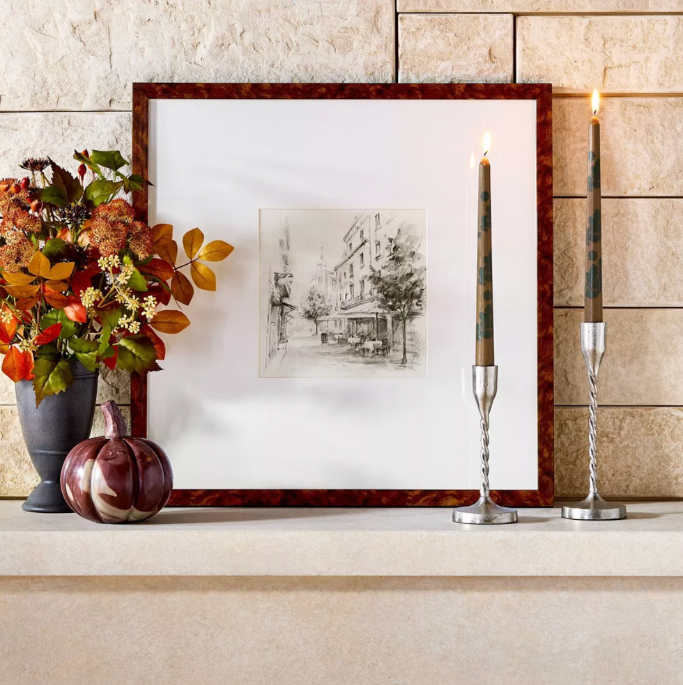 Framed sketch with autumnal decor and lit candles on a stone mantel.