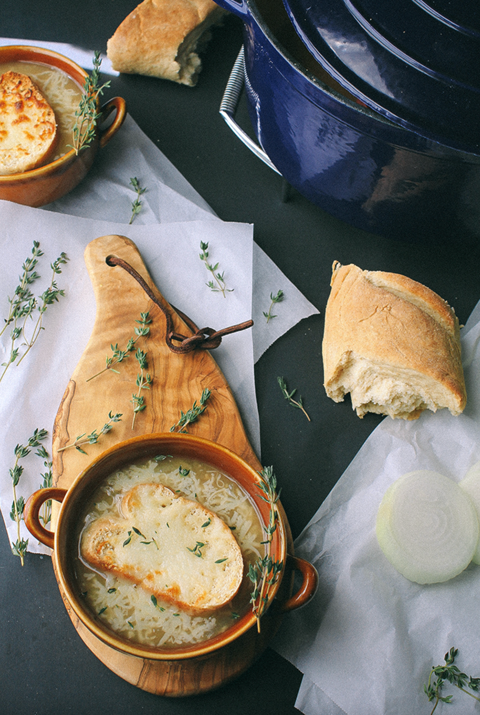 French onion soup with cheese toast, thyme garnish, and bread on a dark table.