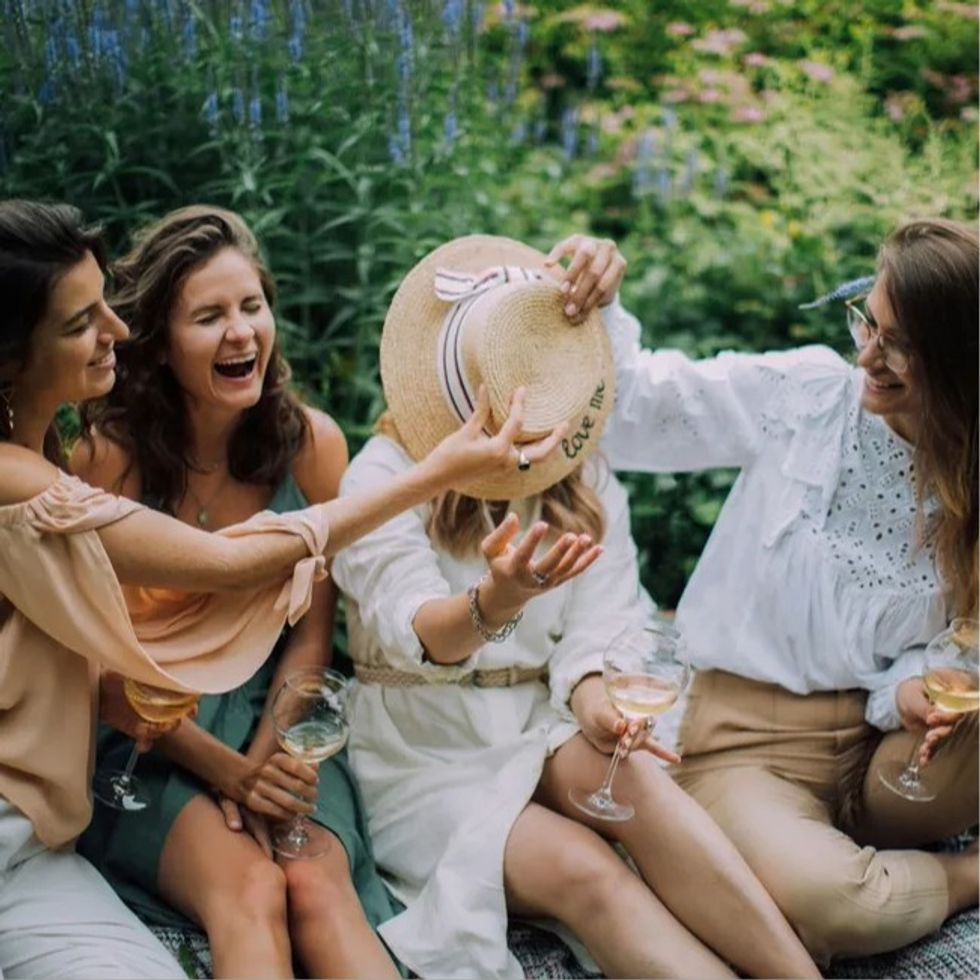 friends laughing and drinking wine while covering their other friend's face with a hat
