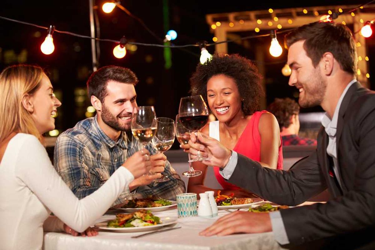 Friends toasting wine glasses at an outdoor dinner under string lights.
