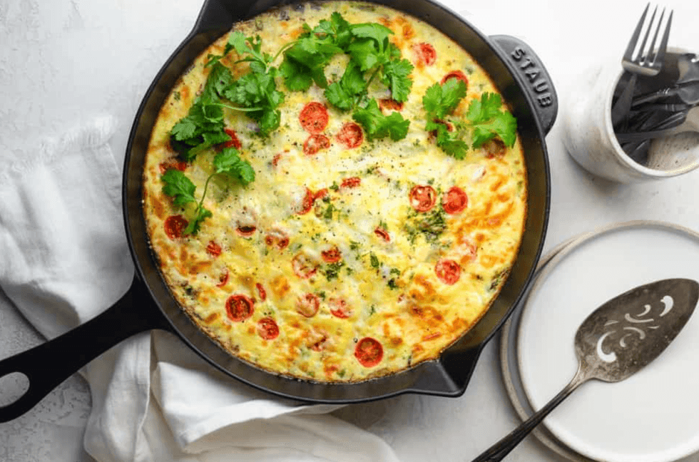 Frittata in a skillet with cherry tomatoes and herbs, accompanied by plates and cutlery.