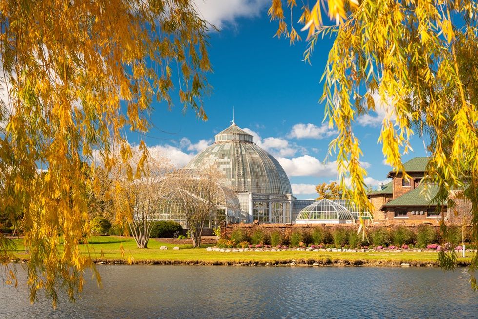 Glass conservatory dome framed by autumn trees and blue sky over a calm pond.
