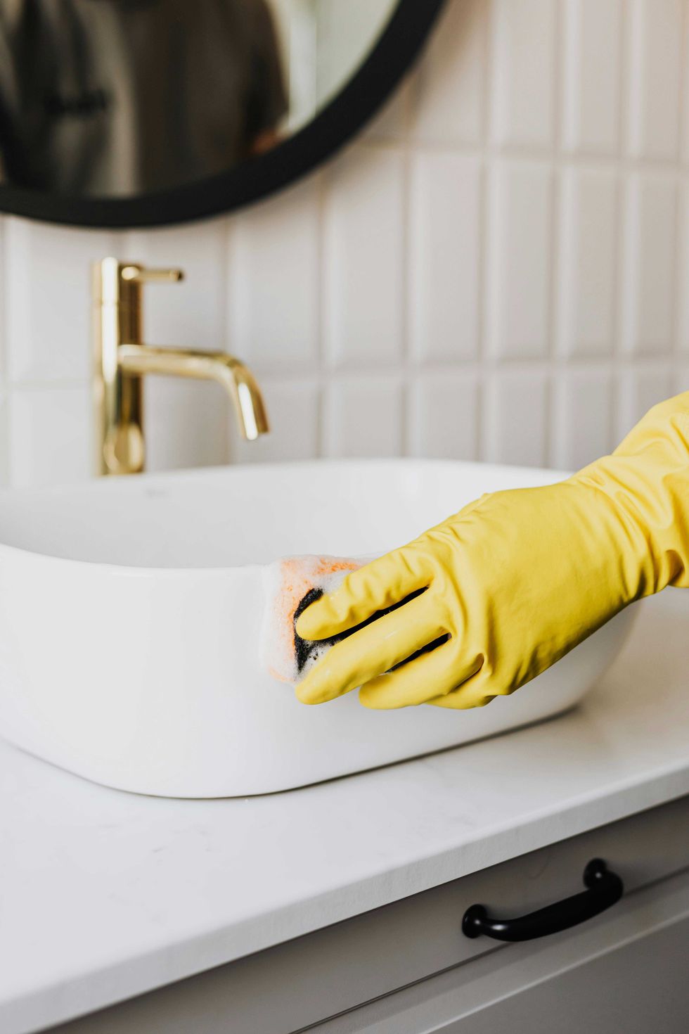 Gloved hand cleaning a white sink with a sponge in a bathroom.