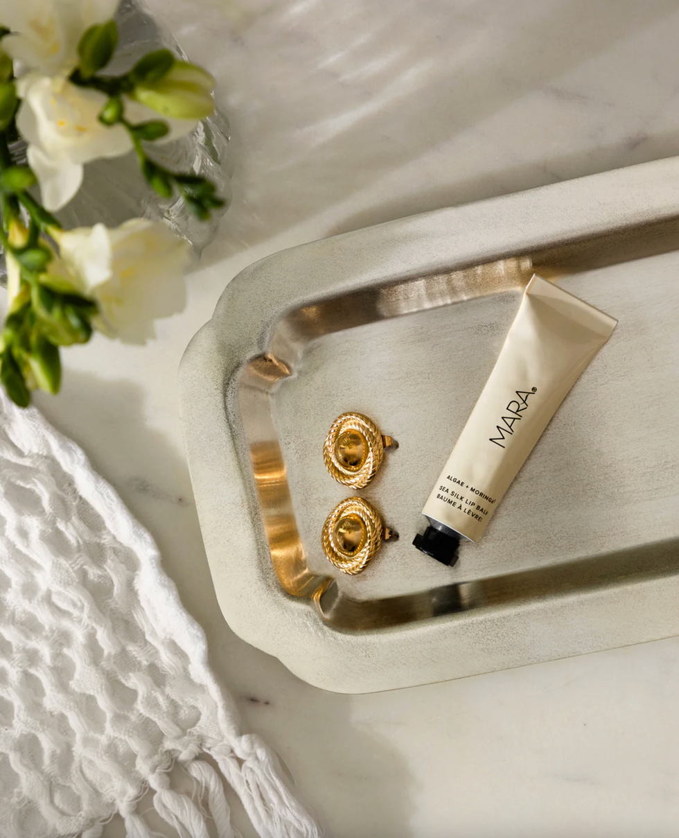 Gold earrings and cream on tray, next to white flowers and cloth.