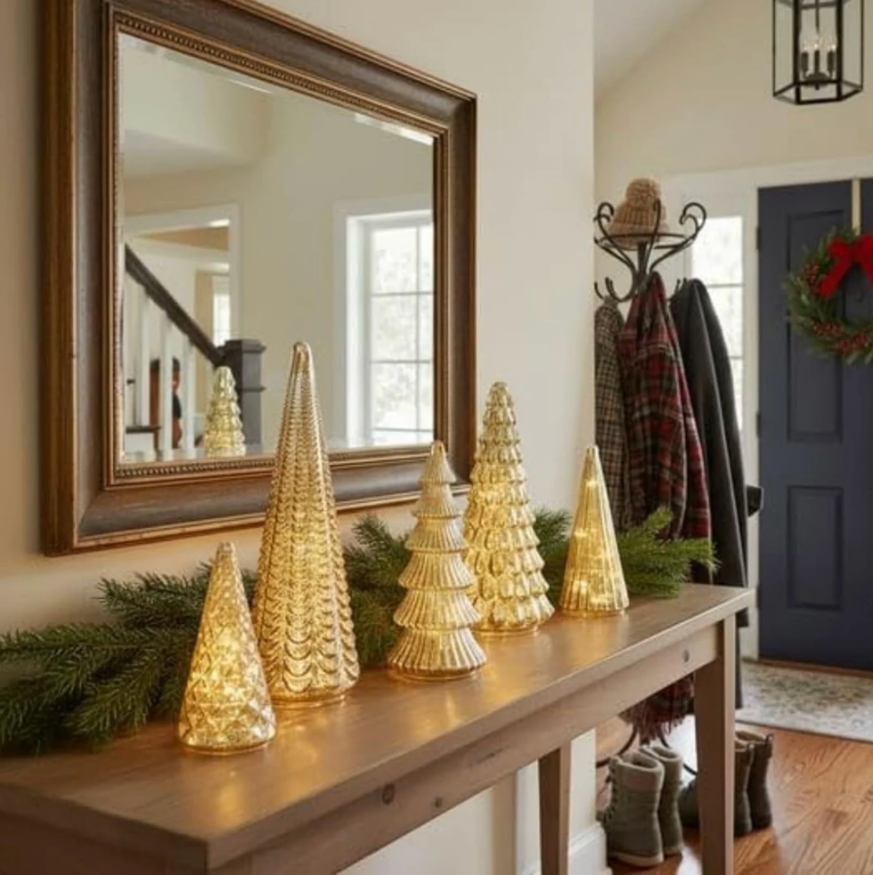 Golden glass trees with lights on a wooden table, mirror, and wreath on door.