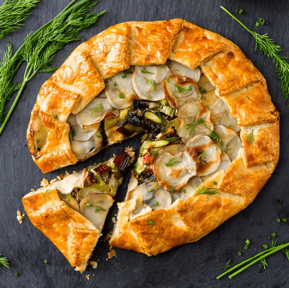 Golden vegetable galette with a slice cut out, garnished with dill, on a dark slate surface.