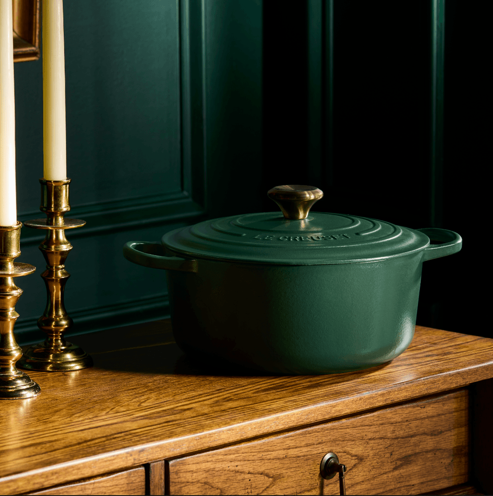 Green cast iron pot on wooden surface, with brass candlesticks, against dark green wall.