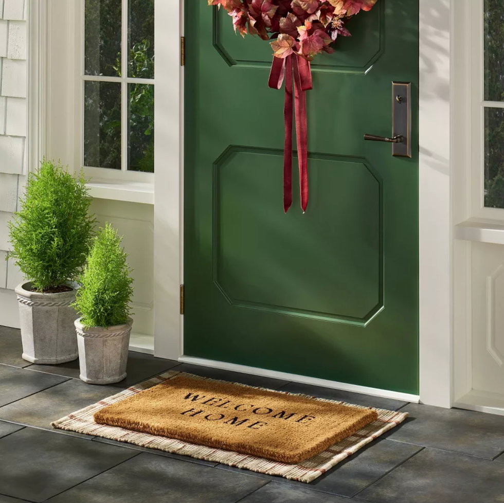 Green door with wreath, welcome mat, and potted plants on a tiled porch.