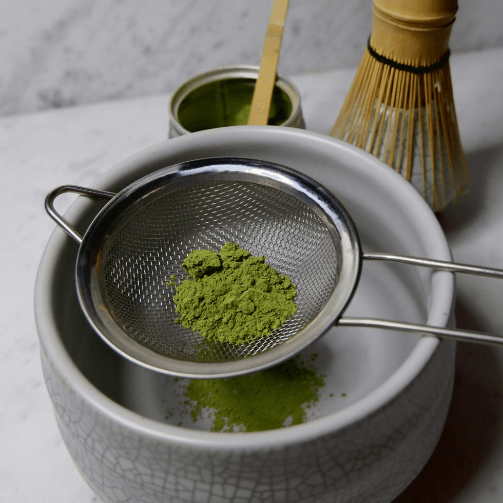 Green matcha powder in a mesh strainer sitting on a small bowl