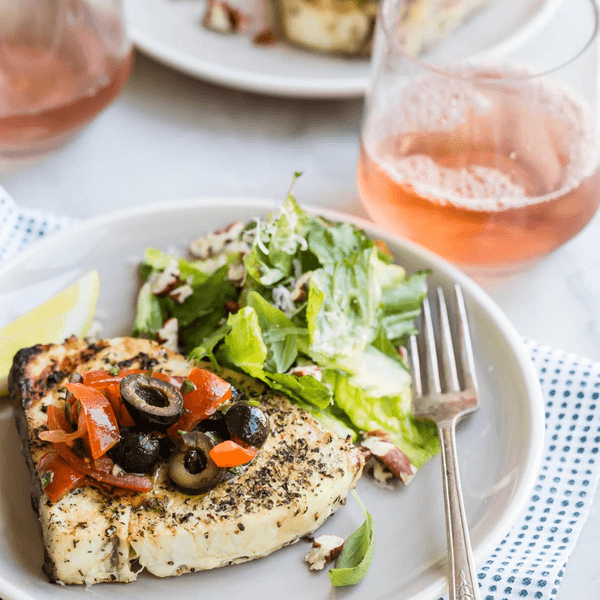 Grilled fish with olives, salad, and a glass of ros\u00e9 wine on a table.