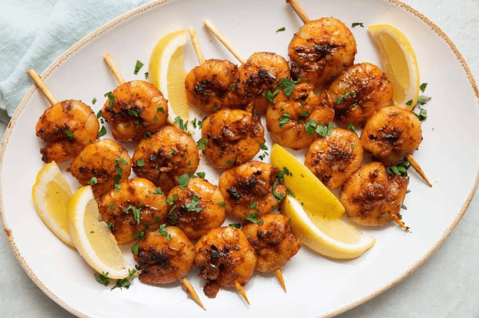 Grilled shrimp skewers with parsley and lemon wedges on a white plate.